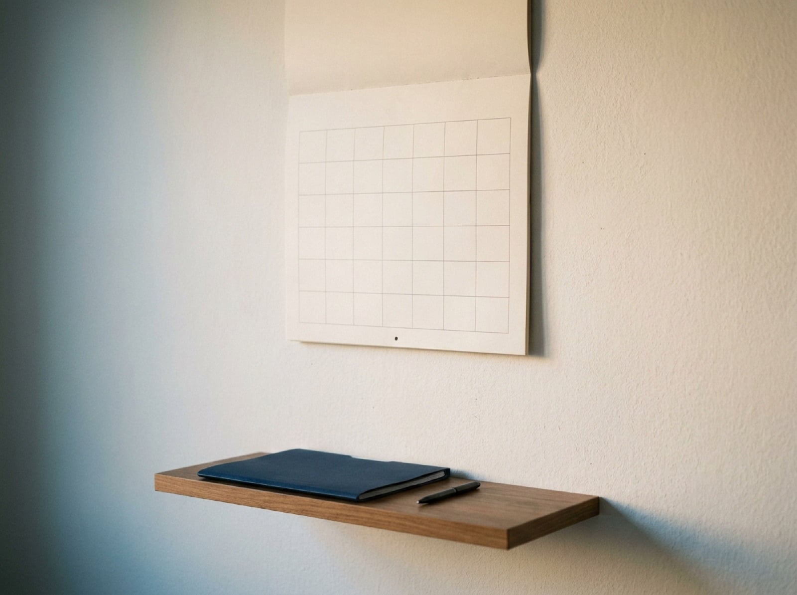 Calendario de pared de mes completo junto a una carpeta cerrada y un bolígrafo sobre una mesa